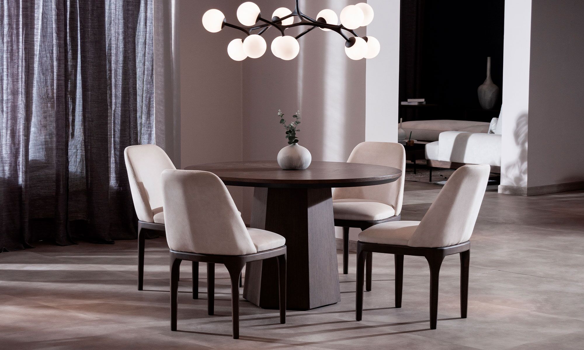 AMBIENTE-COMEDOR-MILANO-REDONDO-4-SILLAS-LISBOA Mesa Comedor Milano redonda con 4 sillas Lisboa de la marca Aristas, vista en diagonal. La mesa tiene una superficie circular, sostenida por una base central robusta en madera color Chocolate. Las sillas Lisboa cuentan con un respaldo elegante, asiento acolchado y acabado en tela suave.